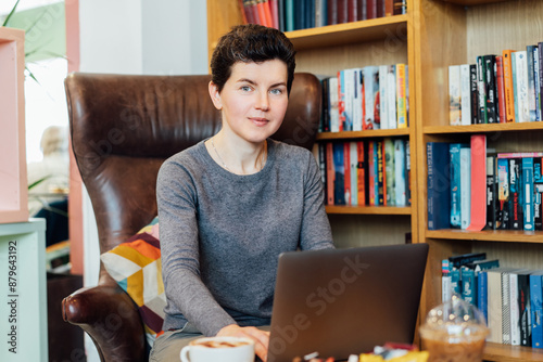 Confident middle aged female expert sits with laptop in leather armchair in cozy office with bookshelf background. 40s middle-age professional portrait of teacher, coach, mentor, therapist, counselor. photo