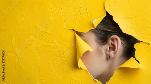 A close-up of a person's ear and hair peeking through a ripped yellow paper background, highlighting textures and colors in a minimalist composition. photo