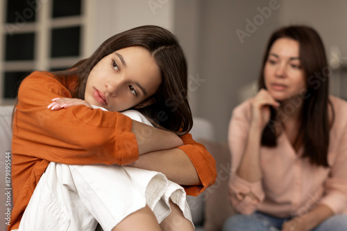 Depressed sad teenage girl sitting and hugging her knees, looking away while mother sitting next daughter. Communication problems between parent and kids photo