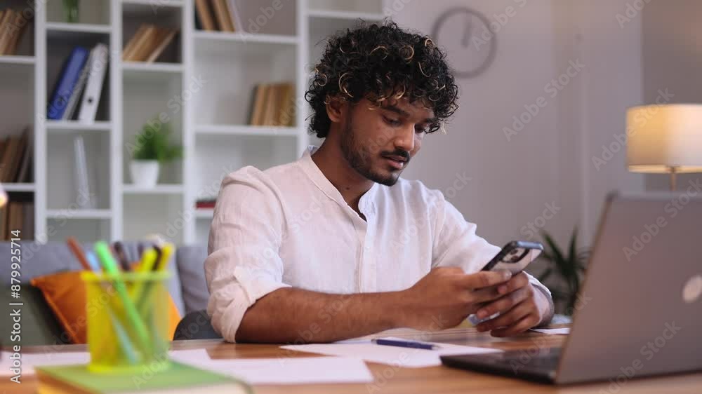 Positive young indian man student relaxing at home take a break from study or work typing browsing products in internet online store reading news or social media profile on smartphone indoors Positive young indian man student relaxing at home take a break from study or work typing browsing products in internet online store reading news or social media profile on smartphone indoors