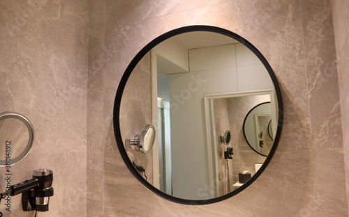 A round bathroom mirror on a tiled wall with view of  a magnifying mirror and hair drier on the wall. Hotel amenities concept.