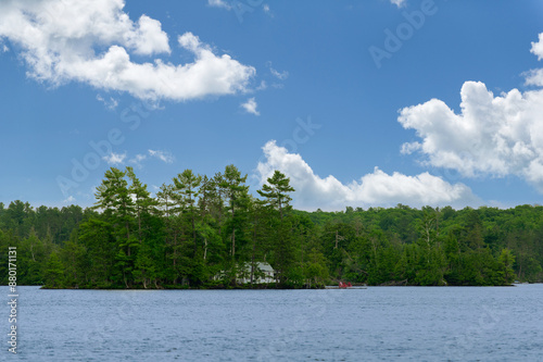 A charming cottage nestled between green trees on a lake in Muskoka, Ontario, Canada. The blue sky reflects in the calm waters, and a dock with red Adirondack chairs adds a pop of color to the serene.