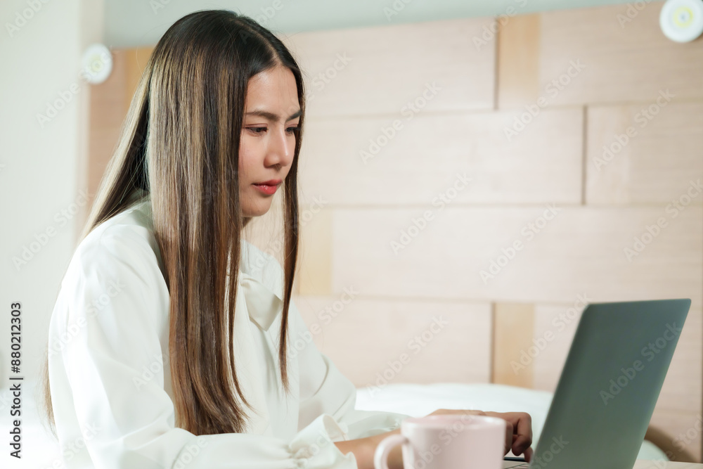 Beautiful Asian woman working from home on holiday, concentrating on laptop with pink cup on table. Cozy bedroom setting with modern lifestyle elements, showcasing remote work and productivity. Beautiful Asian woman working from home on holiday, concentrating on laptop with pink cup on table. Cozy bedroom setting with modern lifestyle elements, showcasing remote work and productivity.