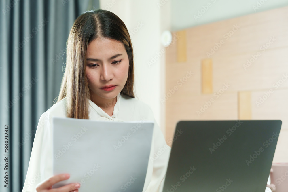 Beautiful Asian woman working from home on holiday, focusing on documents while using laptop, Cozy bedroom setting with modern lifestyle elements, illustrating remote work and productivity. Beautiful Asian woman working from home on holiday, focusing on documents while using laptop, Cozy bedroom setting with modern lifestyle elements, illustrating remote work and productivity.