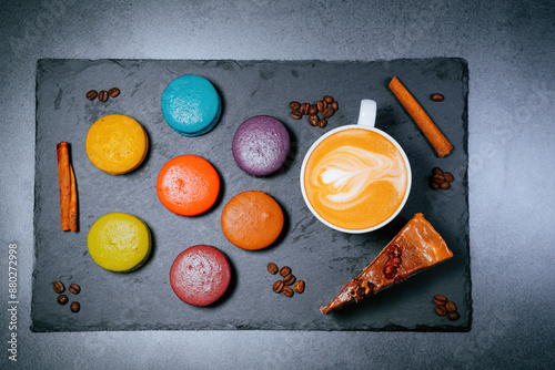 Desserts on a flat black plate. A cup of cappuccino around the sweets of colorful macaroons and carrot cake. Cinnamon sticks and coffee beans. photo