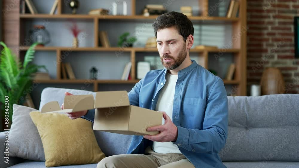 Unhappy man received a parcel with wrong product sitting on sofa in living room at home looking at camera. Bearded male is dissatisfied with shopping in online store. Service issue. Damaged item Unhappy man received a parcel with wrong product sitting on sofa in living room at home looking at camera. Bearded male is dissatisfied with shopping in online store. Service issue. Damaged item