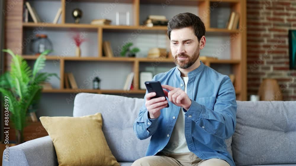 Handsome man using mobile phone sitting on sofa in living room at home. A smiling bearded male browses social networks, does shopping in an online store, chats, writes or reads messages on smartphone Handsome man using mobile phone sitting on sofa in living room at home. A smiling bearded male browses social networks, does shopping in an online store, chats, writes or reads messages on smartphone