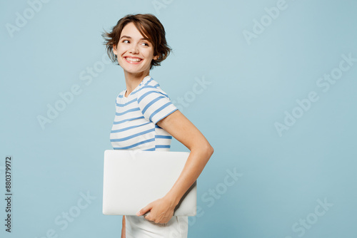 Side view young fun smart IT woman wears striped t-shirt casual clothes hold in hand closed laptop pc computer look aside isolated on plain pastel light blue cyan background studio. Lifestyle concept.