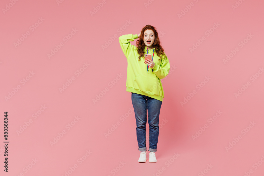 Full body young shocked surprised ginger woman she wears green hoody casual clothes hold head use mobile cell phone isolated on plain pastel light pink background studio portrait. Lifestyle concept. Full body young shocked surprised ginger woman she wears green hoody casual clothes hold head use mobile cell phone isolated on plain pastel light pink background studio portrait. Lifestyle concept.
