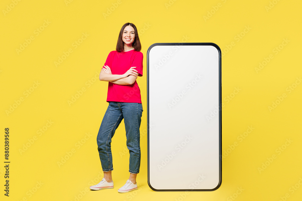 Full body young woman wear pink t-shirt casual clothes stand near big huge blank screen mobile cell phone smartphone with area look camera isolated on plain yellow orange background Lifestyle concept Full body young woman wear pink t-shirt casual clothes stand near big huge blank screen mobile cell phone smartphone with area look camera isolated on plain yellow orange background Lifestyle concept
