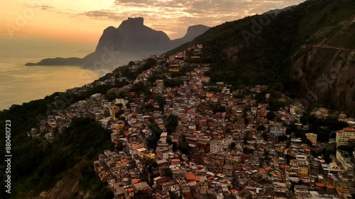 Experience Rio de Janeiro's Favela Vidigal at sunset: a breathtaking aerial drone shot showcasing vibrant houses, the glowing ocean, and majestic Dois Irmaos mountains under warm, golden hues. photo