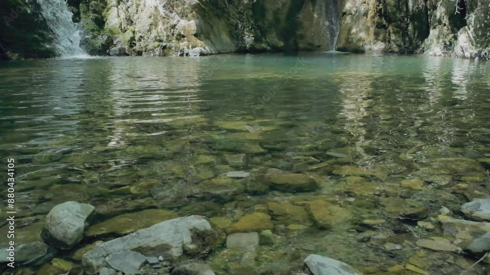 A continuous cascade of water from a distant waterfall feeds into a serene pond, its surface shimmering with hues of green. Slow motion scene revealing ripples and splashes.