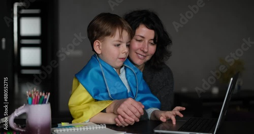 Ukrainian mother and son with flag sitting with laptop at home studying .Happy smiling family hugs and looking at the camera. Ukrainian refugees concept. video footage 4k