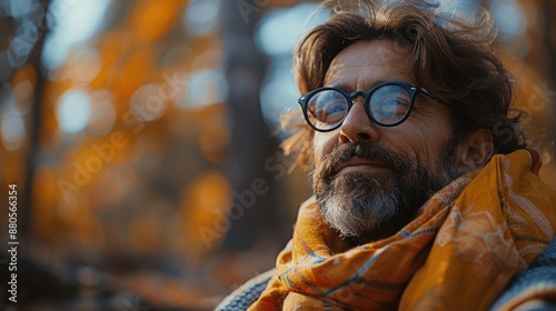 A man in a knit sweater and colorful scarf sitting outdoors amidst autumn foliage, enjoying the serene and crisp fall environment with a calm demeanor.