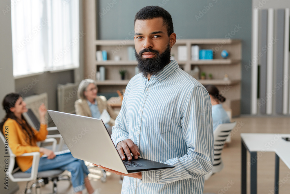African American businessman holding laptop working in office with colleagues looking at camera