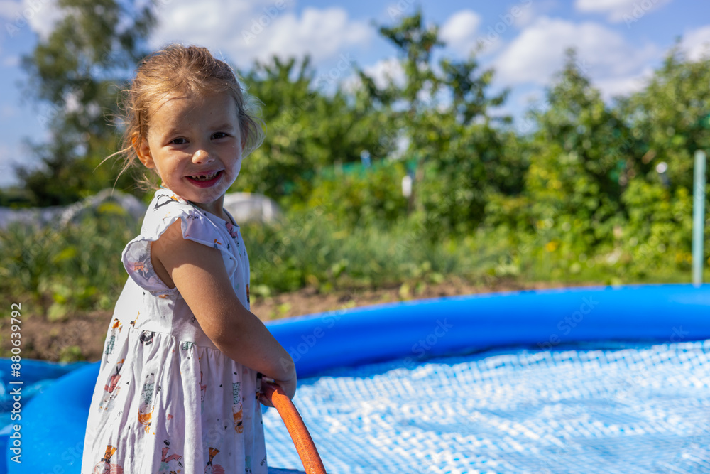 Naklejka premium A beautiful child smiles at the camera and fills an inflatable pool with water from a hose. A cute child is happy to be able to swim in the pool on hot summer days. A child fills a pool.