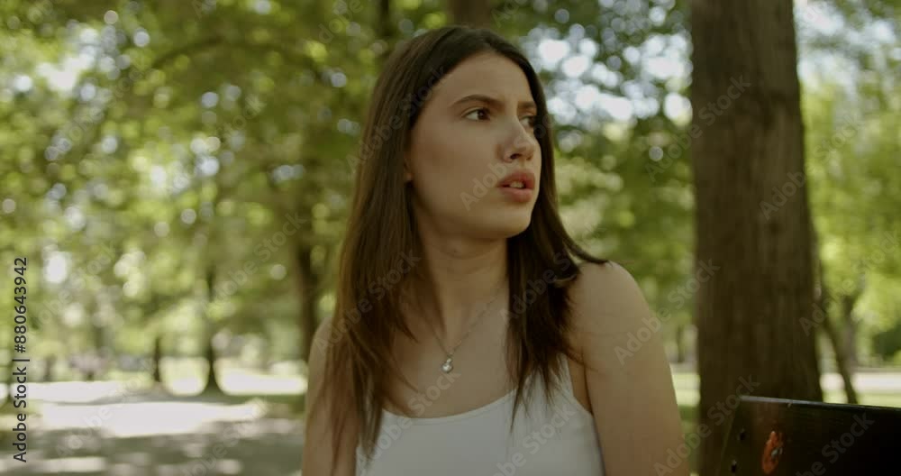 A young woman with a concerned expression sitting on a bench in a park on a sunny day with green trees in the background.