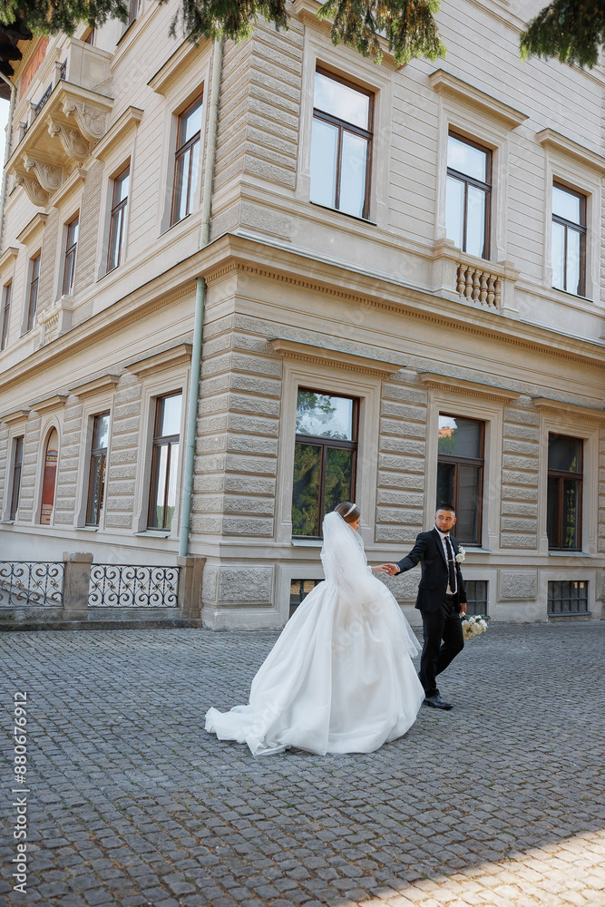 A bride and groom are walking down a cobblestone street in front of a large building. The bride is wearing a white dress and the groom is wearing a black suit. They are holding hands A bride and groom are walking down a cobblestone street in front of a large building. The bride is wearing a white dress and the groom is wearing a black suit. They are holding hands