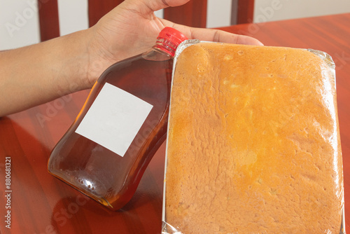 A man holds a bottle of honey with a blank label and a plastic-wrapped mantecada on a wooden table. photo