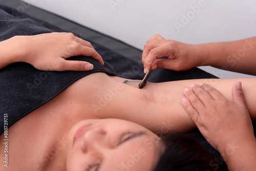 An esthetician applies hot black charcoal wax with a wooden spatula to the armpit of a young woman. Underarm waxing procedure, removing body hair.