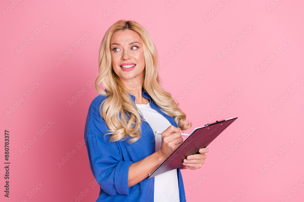 Portrait of optimistic woman with wavy hair dressed blue shirt writing on clipboard look empty space isolated on pink color background Portrait of optimistic woman with wavy hair dressed blue shirt writing on clipboard look empty space isolated on pink color background