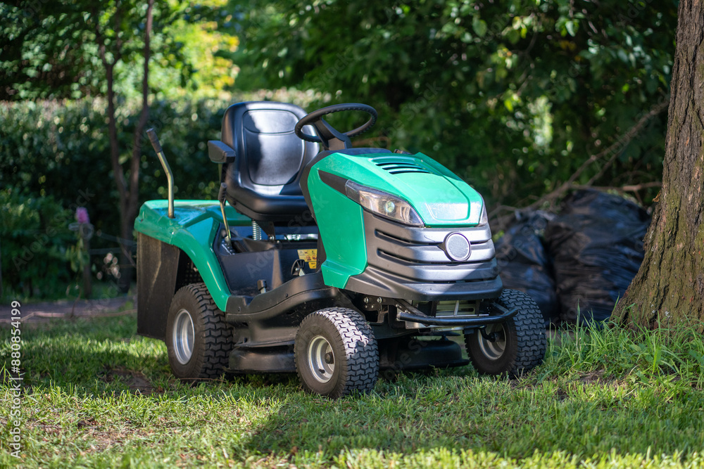 Green contemporary lawnmower on four wheels, located in shade of tree on ground. New turquoise sedentary machine designed to take care of yard near residental house, maintain right level of grass Green contemporary lawnmower on four wheels, located in shade of tree on ground. New turquoise sedentary machine designed to take care of yard near residental house, maintain right level of grass