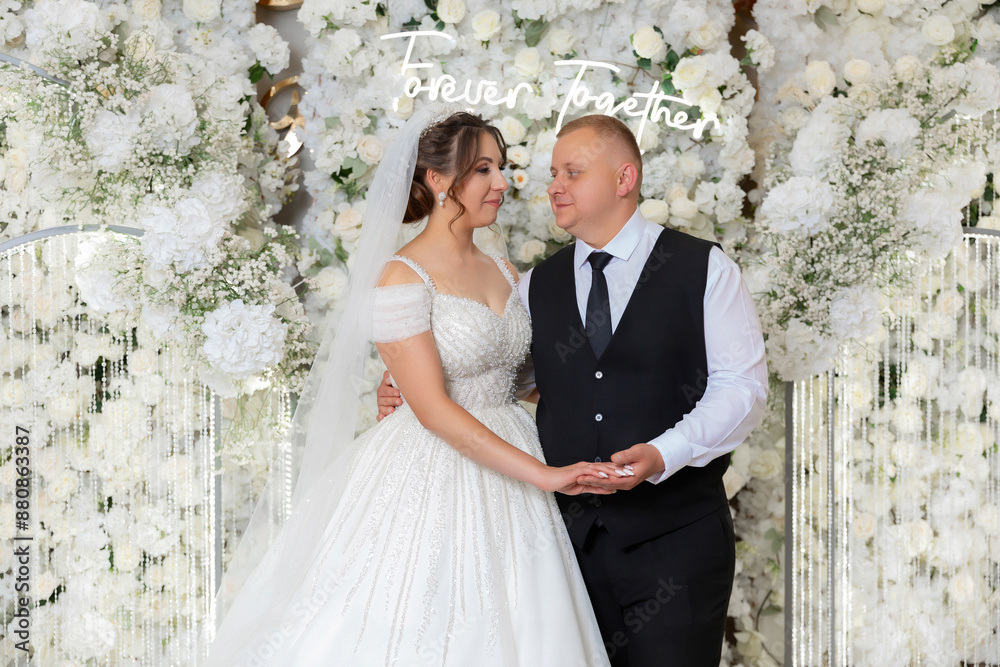 A bride and groom are posing for a picture in front of a white archway. The archway is decorated with flowers and the couple is wearing white and black. Concept of love and happiness A bride and groom are posing for a picture in front of a white archway. The archway is decorated with flowers and the couple is wearing white and black. Concept of love and happiness