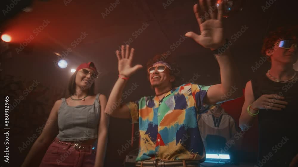 Lowkey shot of charismatic and energetic hipster guy in aviator sunglasses, fancy shirt and with puffy hair dancing in centre of dance floor among trendy ravers to edm in nightclub with flashes