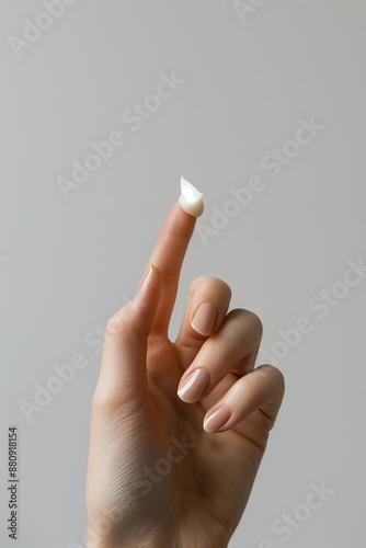 Detailed close-up of a hand with well-manicured nails holding a dab of cream on the fingertip against a neutral background. 