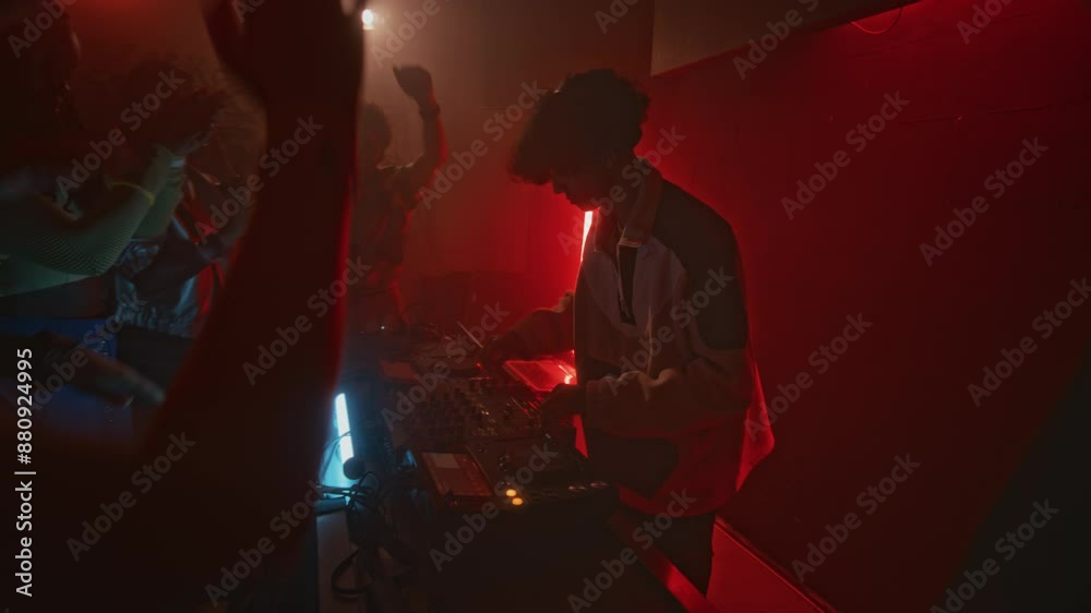 Side shot of cool dj in tracksuit playing electronic dance music on audio mixer to raving crowd during retro party in red neon lit nightclub