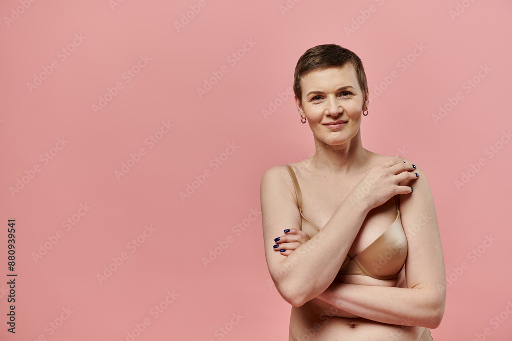 A woman with short hair and bra smiles confidently against a pink background. A woman with short hair and bra smiles confidently against a pink background.