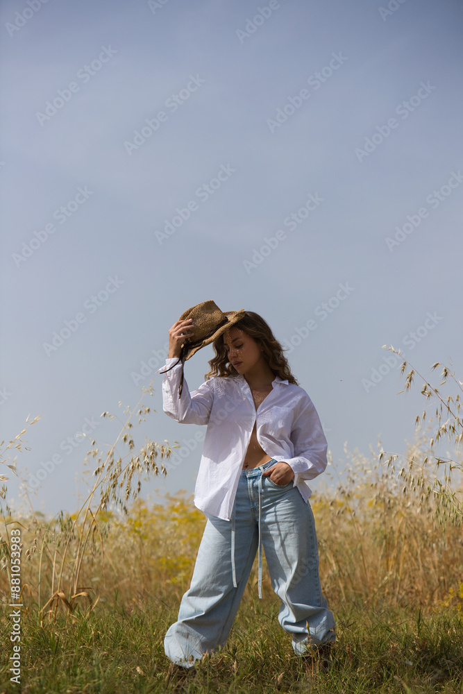 pretty young blonde woman with light eyes is dressed in white shirt, jeans, boots and hat standing in a field planted with oats. The farmer is posing for the photo, in the background the blue sky.