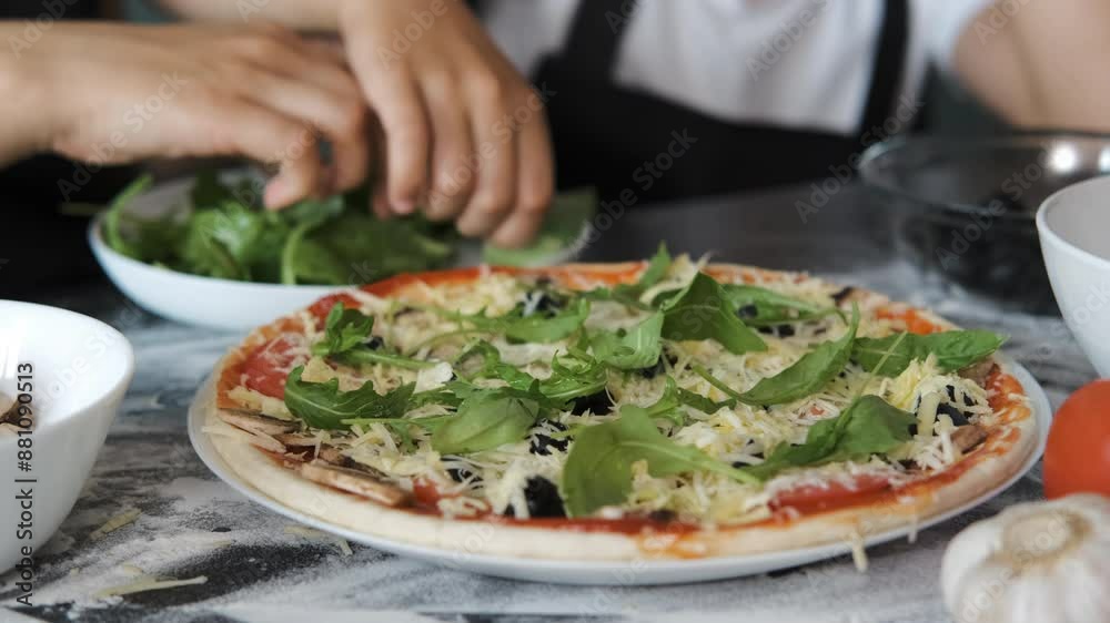 Close-up of mother and daughter preparing a tasty homemade vegan pizza, adding arugula to the pizza crust, slow motion. Family lifestyle concept