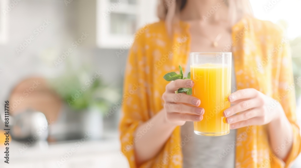 Fototapeta premium Woman in a yellow robe holding a glass of orange juice in a bright kitchen, capturing the essence of a fresh and healthy morning routine.