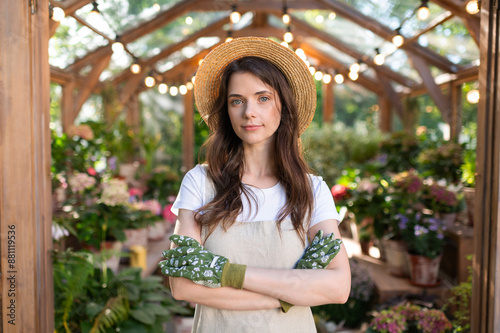 Fotografie Young woman in a straw hat and apron standing in a greenhouse with crossed arms,