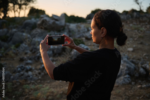 Woman taking photo with smartphone in a rocky outdoor setting during sunset