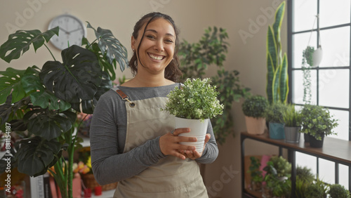 Billede på lærred A smiling hispanic woman holding a potted plant indoors at a flower shop surrounded by greenery