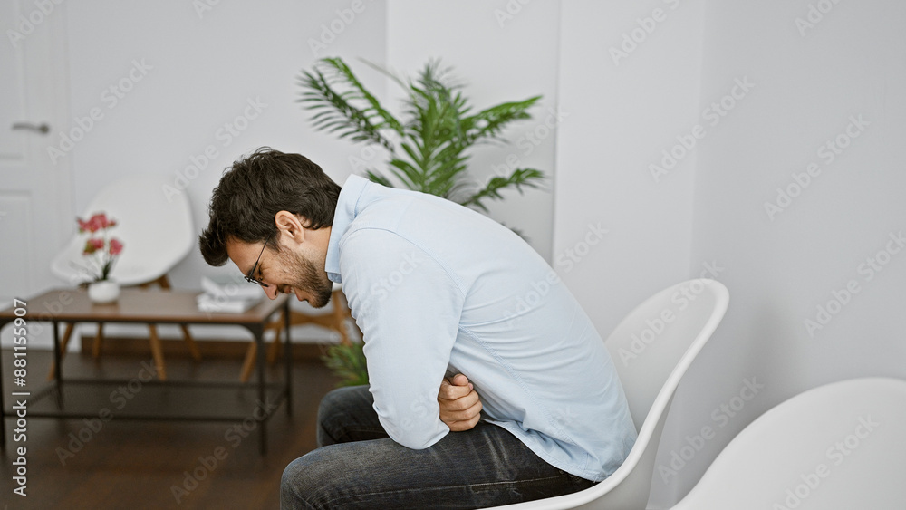 A pained young hispanic man clutches his stomach in a modernly furnished white waiting room, evoking a sense of urgency. A pained young hispanic man clutches his stomach in a modernly furnished white waiting room, evoking a sense of urgency.