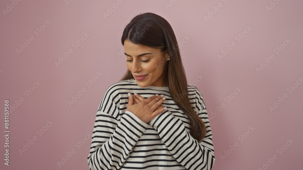 Fototapeta premium Serene hispanic woman with hands on chest, standing against a pink wall, displaying a gesture of gratitude.