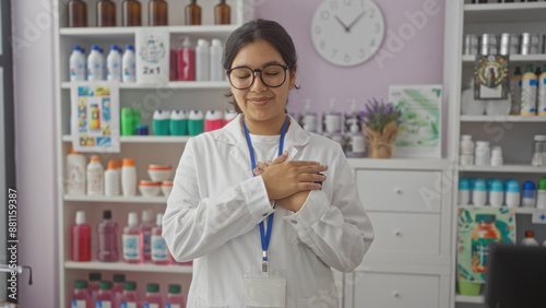 A young hispanic brunette woman in a white coat, standing indoors at a pharmacy, smiling with her hands clasped over her chest.