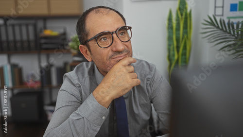 Middle-aged hispanic man in an office, wearing glasses and deep in thought, with shelves and plants in the background.