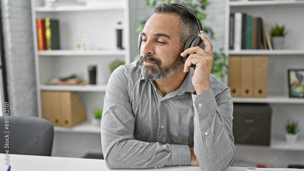 Handsome mature hispanic man with grey beard using headphones in a modern office interior.