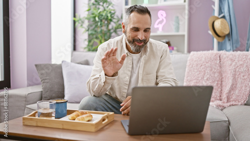 A cheerful mature man with a beard and grey hair video calling from home, using a laptop on a sofa with a neon sign in the background.