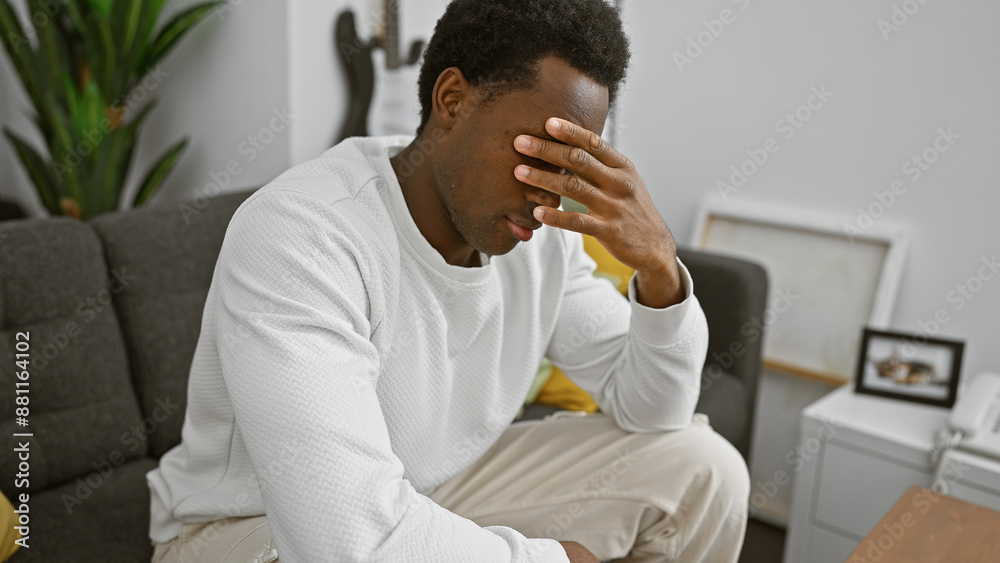 A distressed young african american man sitting indoors in a modern living room covering his face with his hand. A distressed young african american man sitting indoors in a modern living room covering his face with his hand.