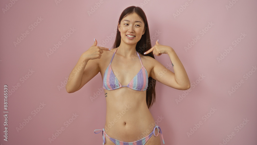 Naklejka premium Beautiful young chinese woman in a colorful bikini smiling and pointing at herself, isolated against a pink background, showcasing her confidence and charm.