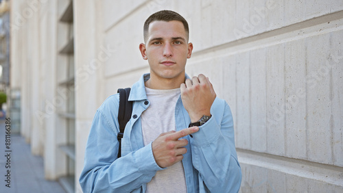 Hispanic young man in casual attire pointing at his watch on a city street, looking punctual and ready.