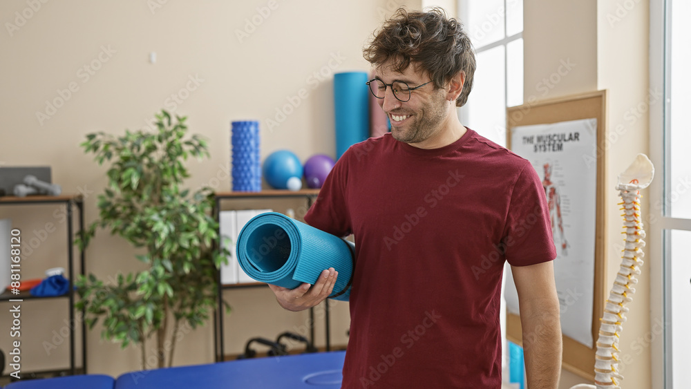 Fototapeta premium Smiling young hispanic man holding a yoga mat in a bright rehab clinic room with physiotherapy equipment.