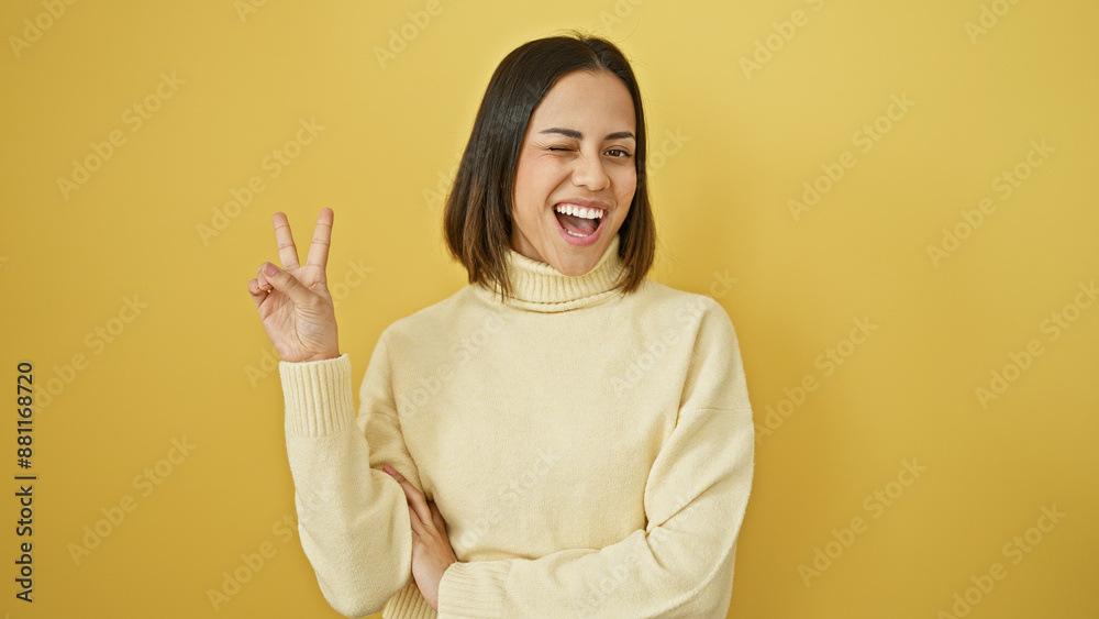 A cheerful young hispanic woman making a peace sign against a plain yellow background A cheerful young hispanic woman making a peace sign against a plain yellow background