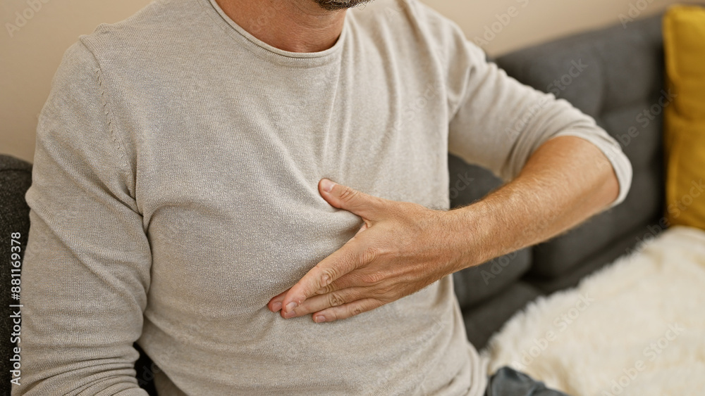 A middle-aged hispanic man clutching his chest in pain while sitting in a living room at home. A middle-aged hispanic man clutching his chest in pain while sitting in a living room at home.