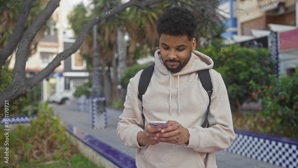 African american man using smartphone while walking in a city park with trees and a tiled bench. African american man using smartphone while walking in a city park with trees and a tiled bench.